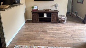 Un soggiorno con pavimento in legno, una TV a schermo piatto montata a parete, un tavolino in legno con un telecomando e delle carte da gioco, e un tappeto decorato.