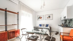 A compact, modern room with a glass dining table, two brown chairs, a sofa with blue cushions, a bed, a kitchenette, and a large window with sheer curtains. - Apartment with balcony near Maradona Stadium (Napoli)