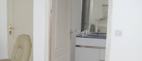 Una habitación con una silla color beige, una puerta blanca y un baño con lavamanos y espejo.