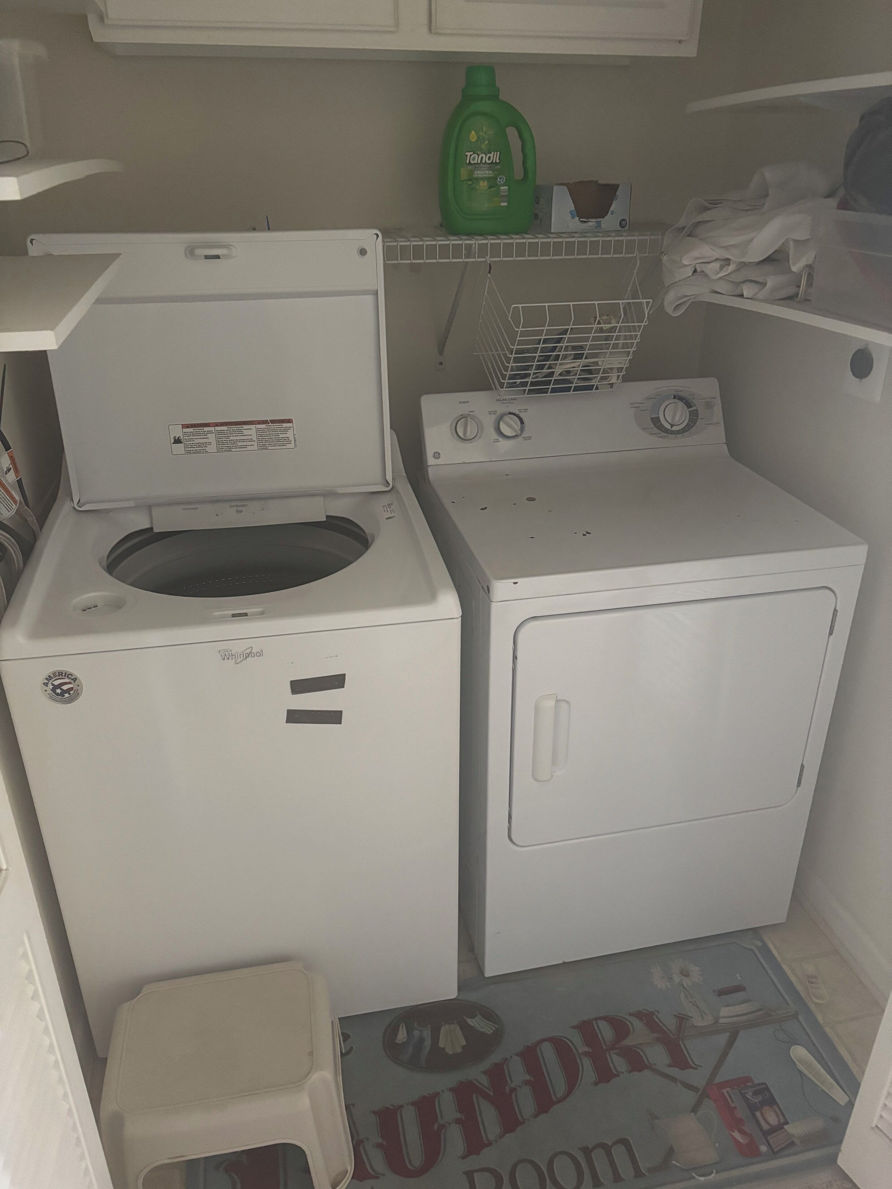 A compact laundry room with a Whirlpool washer and dryer, a small stool, and a shelf with a detergent bottle.