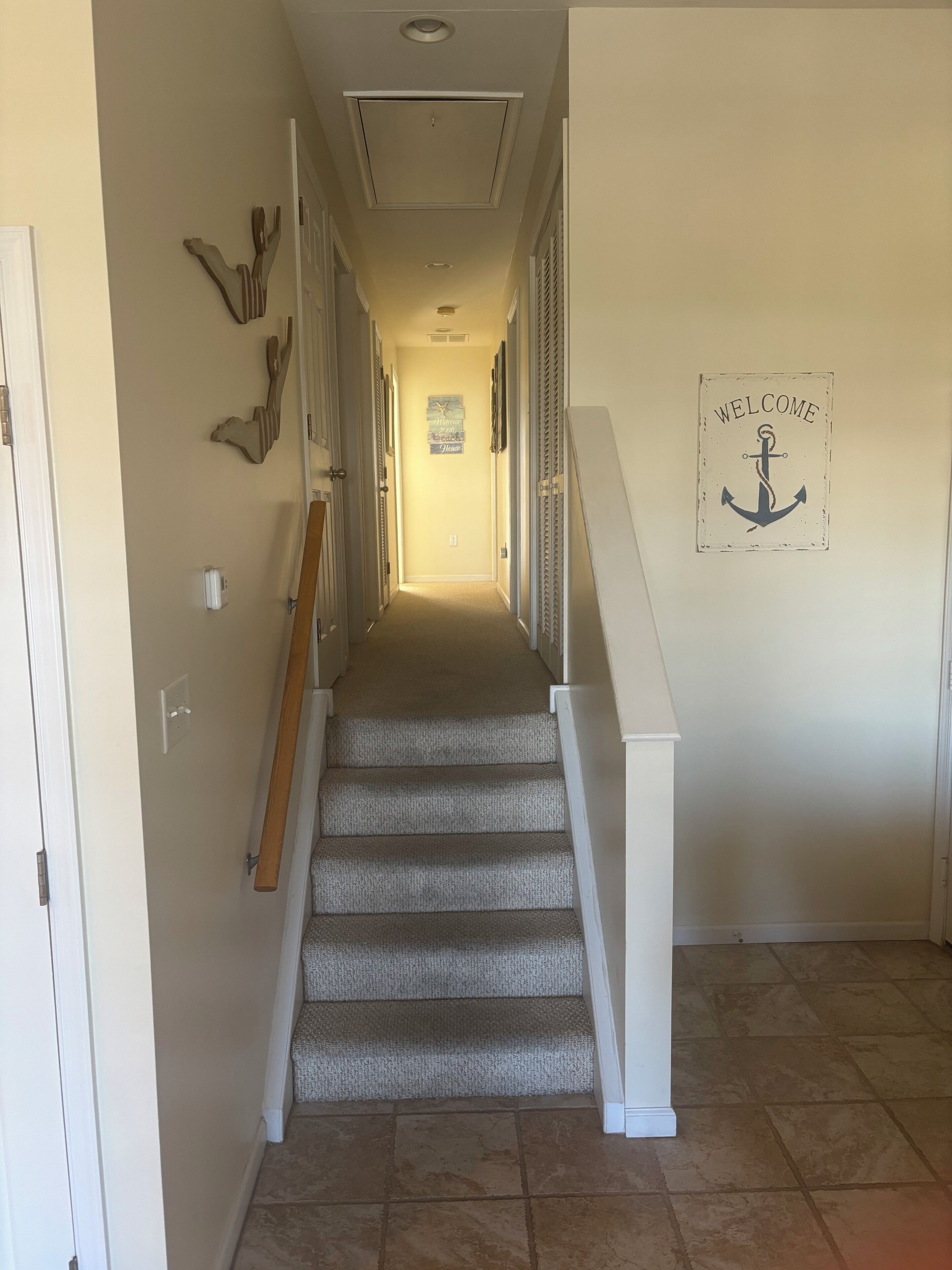 A hallway with carpeted stairs, a welcome sign with an anchor, and a wall-mounted light fixture.