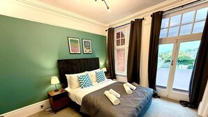 A neatly made bed with a dark bedspread and two pillows, a bedside table with a lamp, and a window with curtains. - Boutique King Balcony Room (Southwold)