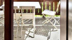 Eine Holzdielen-Terrasse mit weißen Metallstühlen und einem Tisch, mit Blick auf eine Reihe von Gebäuden mit geneigten Dächern und einer Bergkulisse.