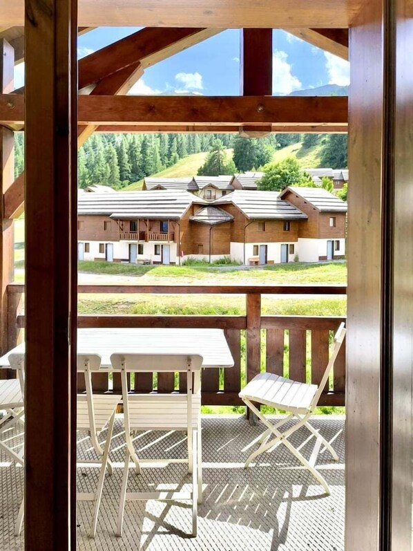 A wooden deck with white metal chairs and a table, overlooking a row of buildings with sloped roofs and a mountainous backdrop.