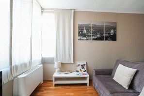 A modern living room with a white radiator, a grey sofa, a white side table with a lamp, and a framed cityscape picture on the wall. - Torino | Nizza Apartment   (Torino)