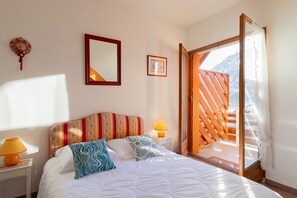 A bedroom with a bed, a nightstand with a lamp, a mirror, and a picture on the wall. There is a window with a view of a mountain. - Le Parc Neige - Vue Montagne (Briancon)