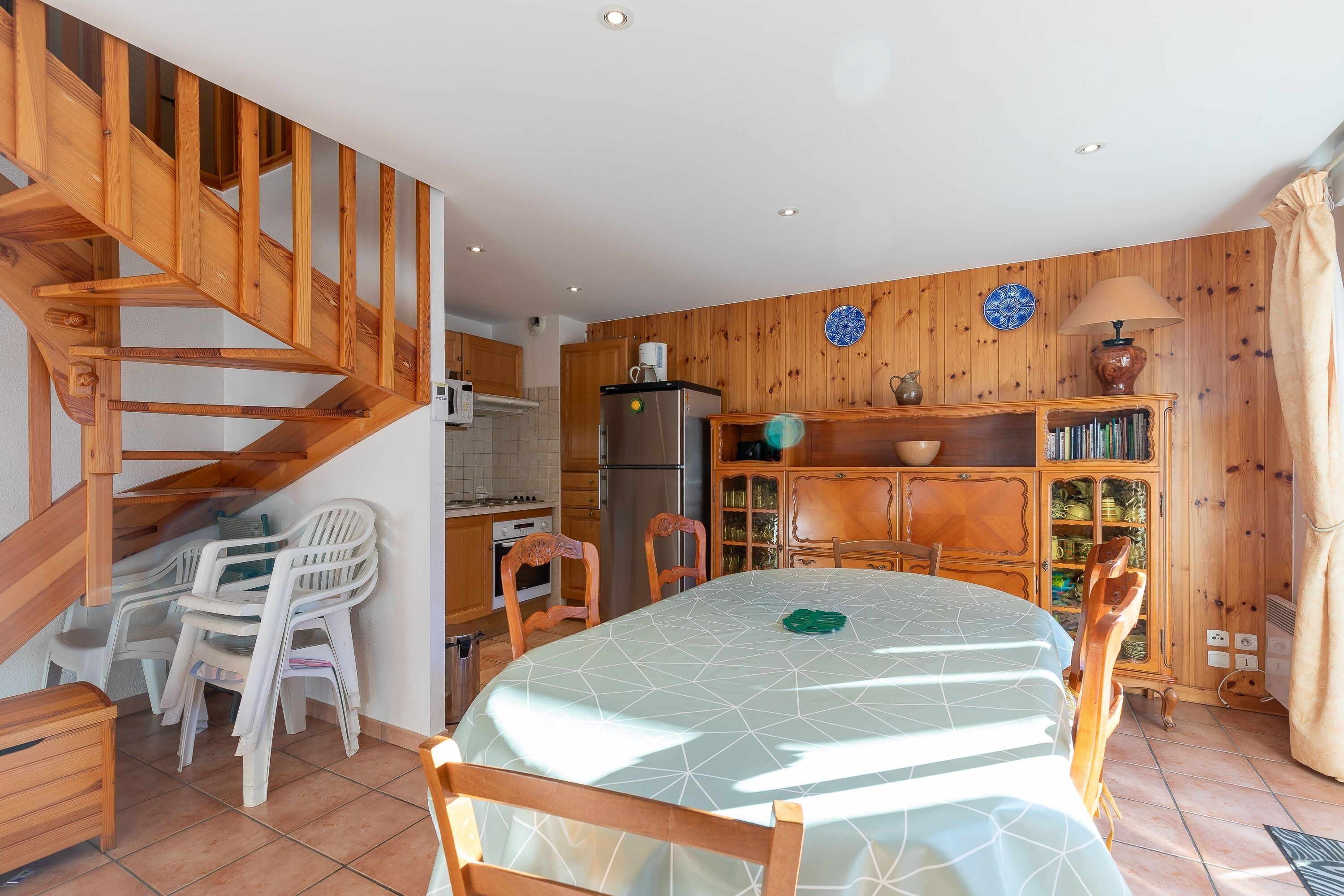 Een eetgedeelte met een tafel gedekt voor vier personen, houten stoelen, een trap, een keuken met houten kastjes en een koelkast.
