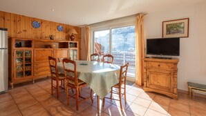 A dining area with a table and chairs, a wooden cabinet with glass doors, a television, and a window with curtains. - Le Parc Neige - Vue Montagne (Briancon)