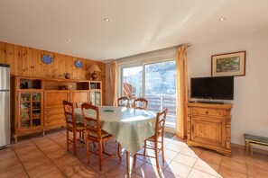 A dining area with a table and chairs, a wooden cabinet with glass doors, a television, and a window with curtains. - Le Parc Neige - Vue Montagne (Briancon)