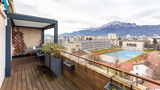 Un balcone con pavimento in legno, un tavolo, sedie e piante in vaso. Sullo sfondo, si vede una scuola con un campo sportivo e degli edifici.