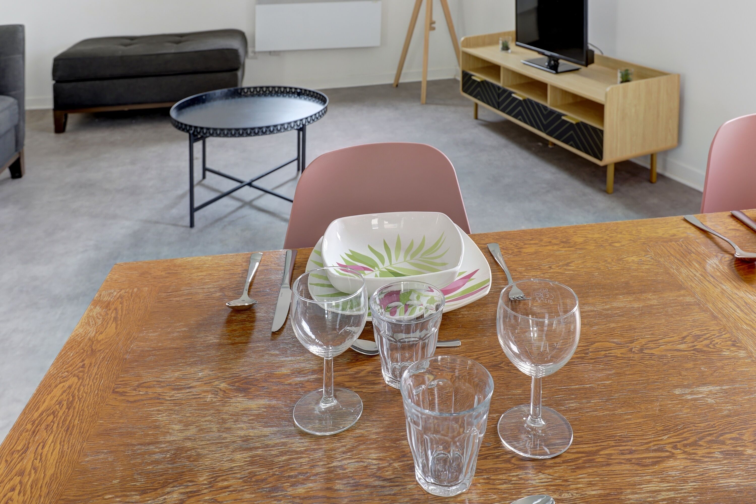 Un comedor con una mesa de madera, cuatro copas de vino, un plato con diseño de hoja verde y una mesita redonda con estructura de metal negro.