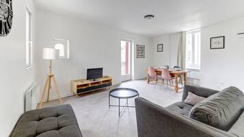 Una sala de estar moderna con un sofá gris, una mesa de centro redonda, un televisor sobre un mueble de madera, un rincón comedor con una mesa y sillas, y un amplio ventanal que proporciona luz natural.