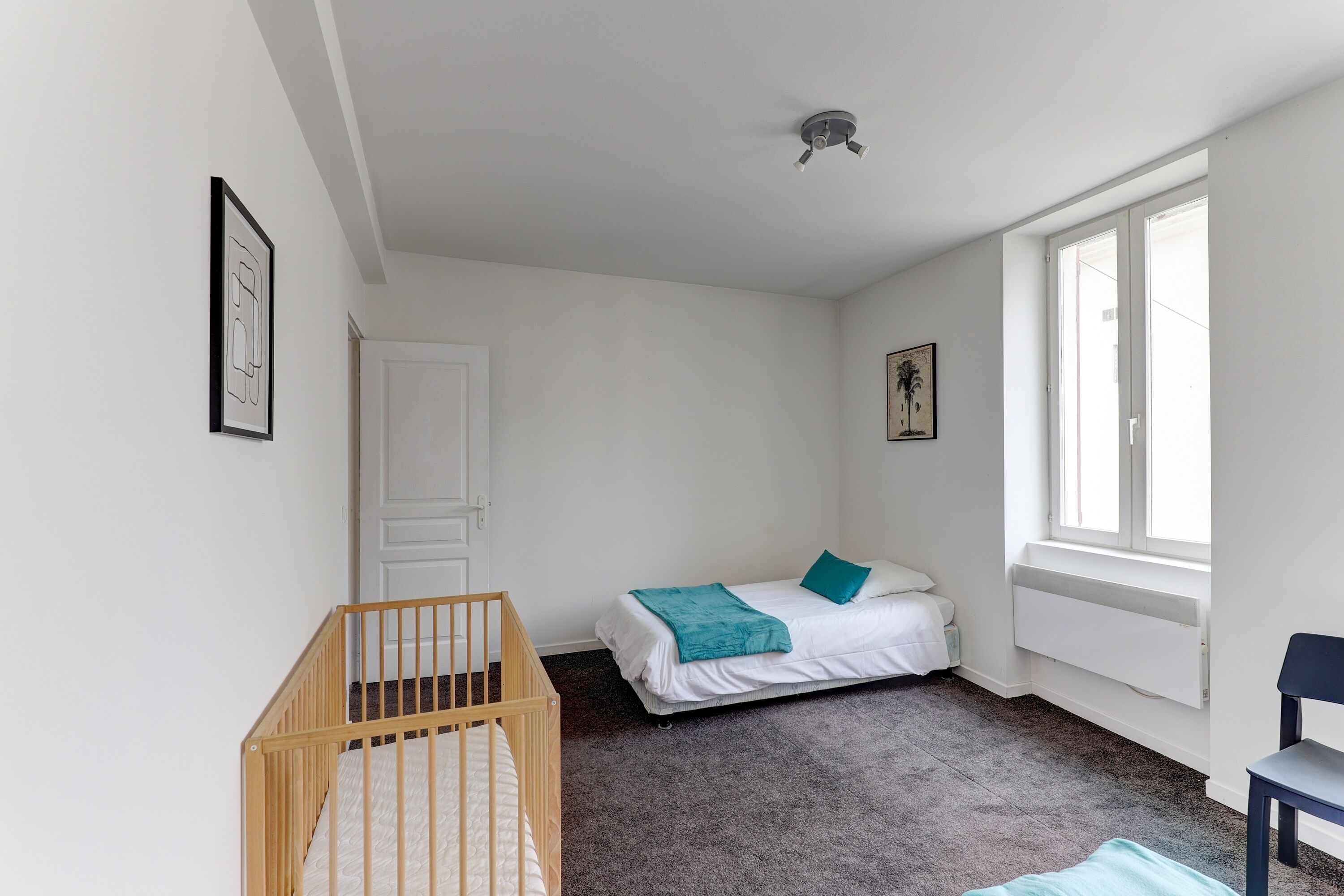 Een kamer met een wieg, een bed met een blauwgroene deken, een stoel en een raam met uitzicht.