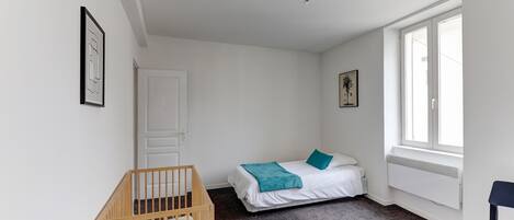 Een kamer met een wieg, een bed met een blauwgroene deken, een stoel en een raam met uitzicht.