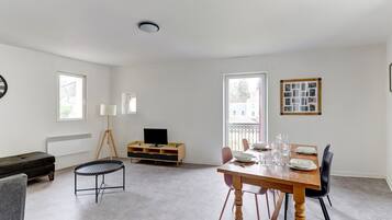 Una sala de estar moderna con una mesa de comedor preparada para cuatro personas, un sofá, una pequeña mesa de centro y un televisor sobre un mueble de madera.