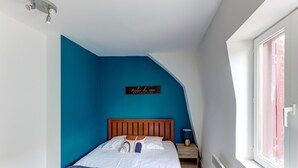 Une chambre avec un plafond en pente, un lit doté d’une tête de lit en bois, une table de chevet avec une lampe et une fenêtre donnant sur un bâtiment rouge.