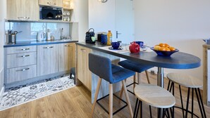 A modern kitchen with wooden cabinets, a microwave, and a dining area with blue chairs and stools. - Les Camélias - 5min à pied du Port (Courseulles-sur-Mer)