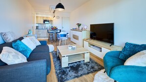 A modern living room with a sofa, a coffee table, and a TV. There is a kitchen in the background with a dining area. - Les Camélias - 5min à pied du Port (Courseulles-sur-Mer)