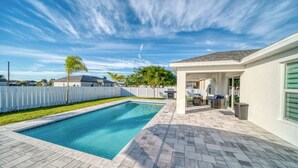 Una piscina rectangular con una solana empavedada, un patio cubierto y una cerca blanca que delimita la propiedad.