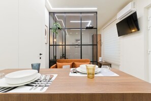 Un comedor con una mesa de madera dispuesta para dos personas, un televisor montado en la pared y un ventanal corredero que da a la cocina.