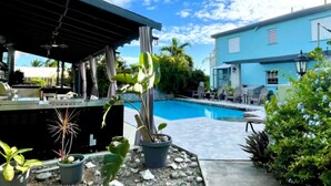 Zona de piscina con bar cubierto, plantas en macetas y un edificio azul al fondo.