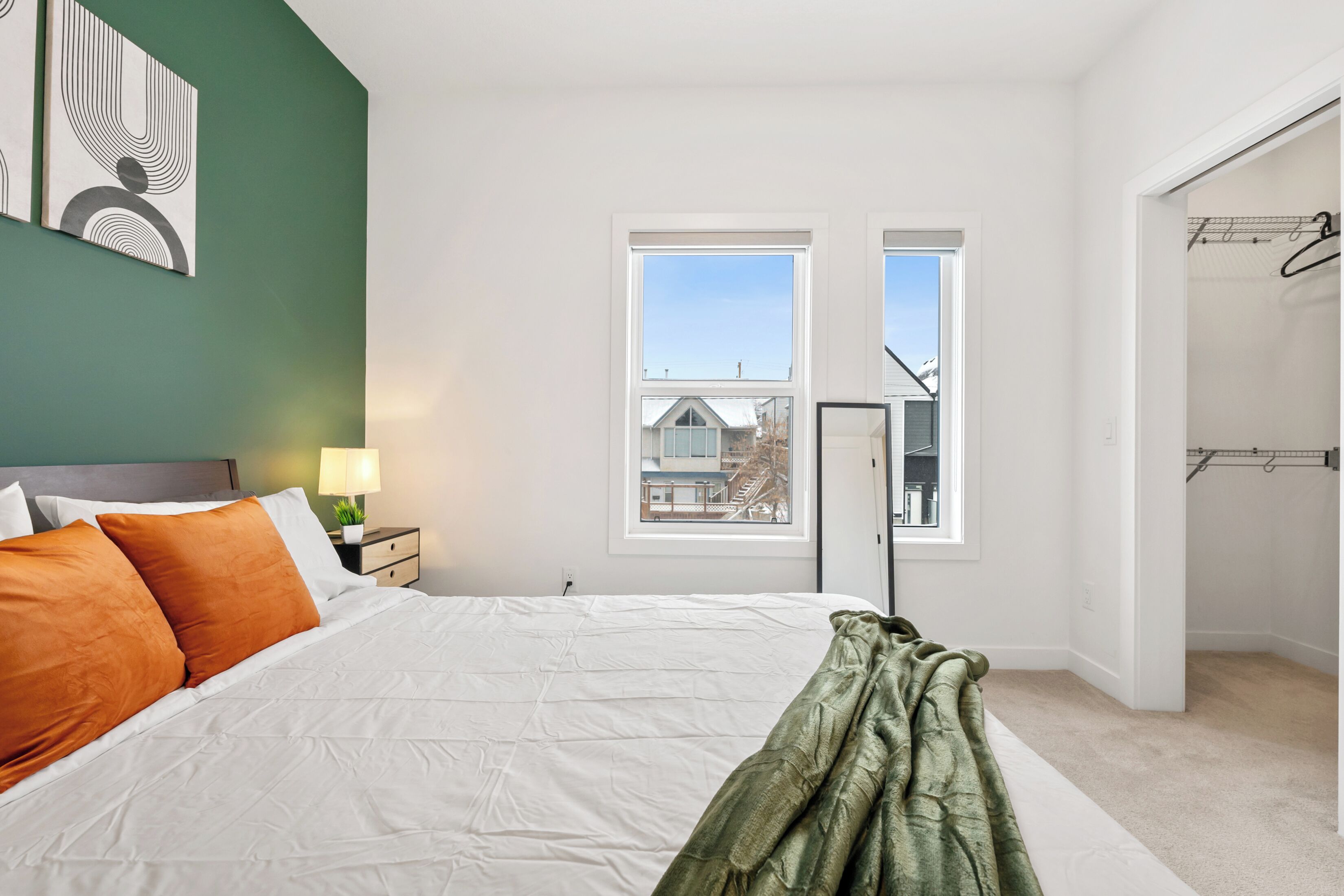 Une chambre à coucher avec un lit, une table de chevet, une lampe et une fenêtre donnant sur une maison.