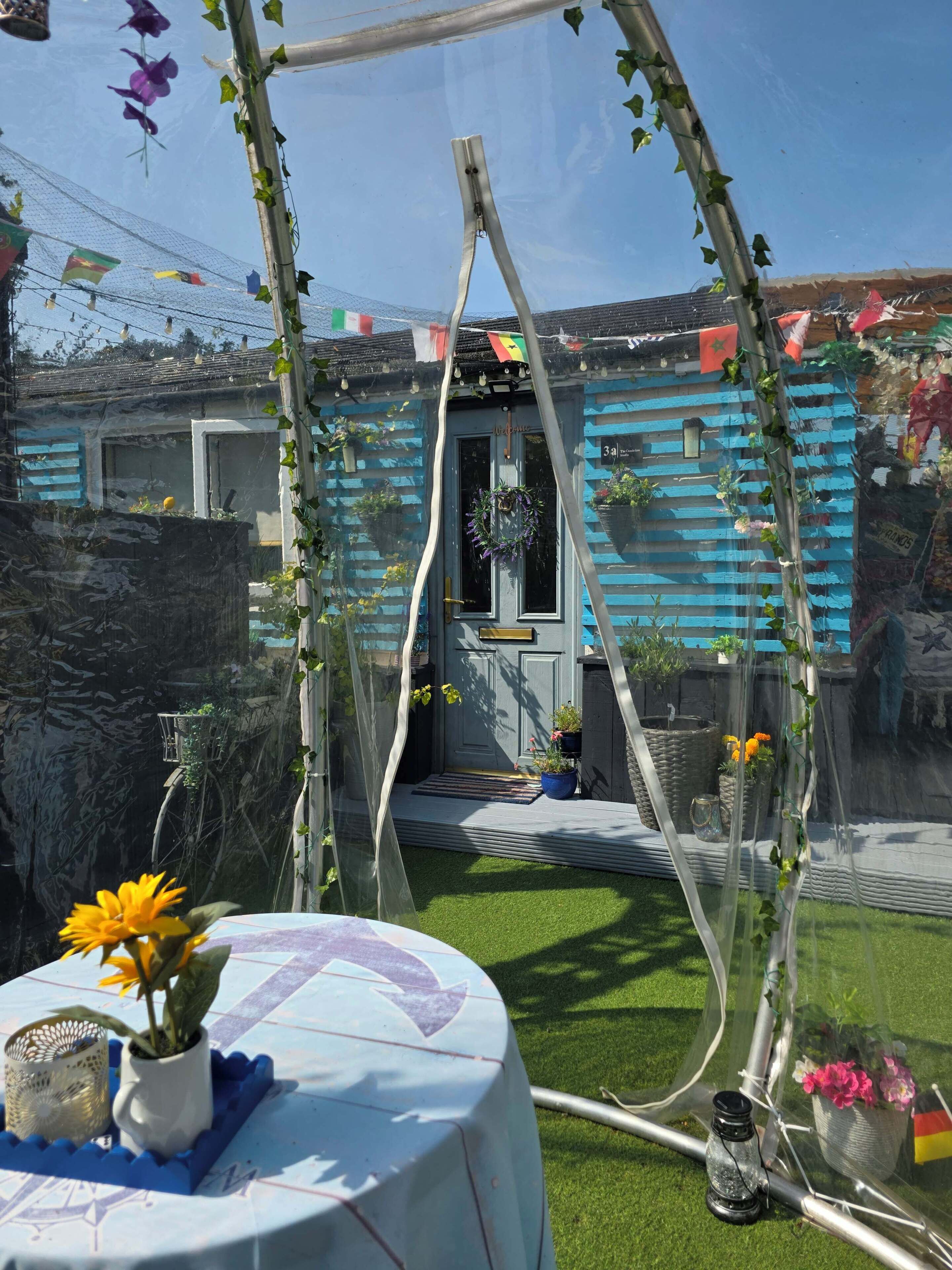 Un jardín con un edificio azul, una pérgola cubierta de plantas trepadoras y una mesa con una planta en maceta.