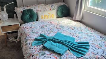 Un lit bien fait, recouvert d’une couvre-lit à motifs floraux, un oreiller orné du mot « sourire », une table de chevet avec un ventilateur, et un mur sur lequel est accroché un panneau avec l’inscription « DES CÂLINS ET DES BISOUS ».
