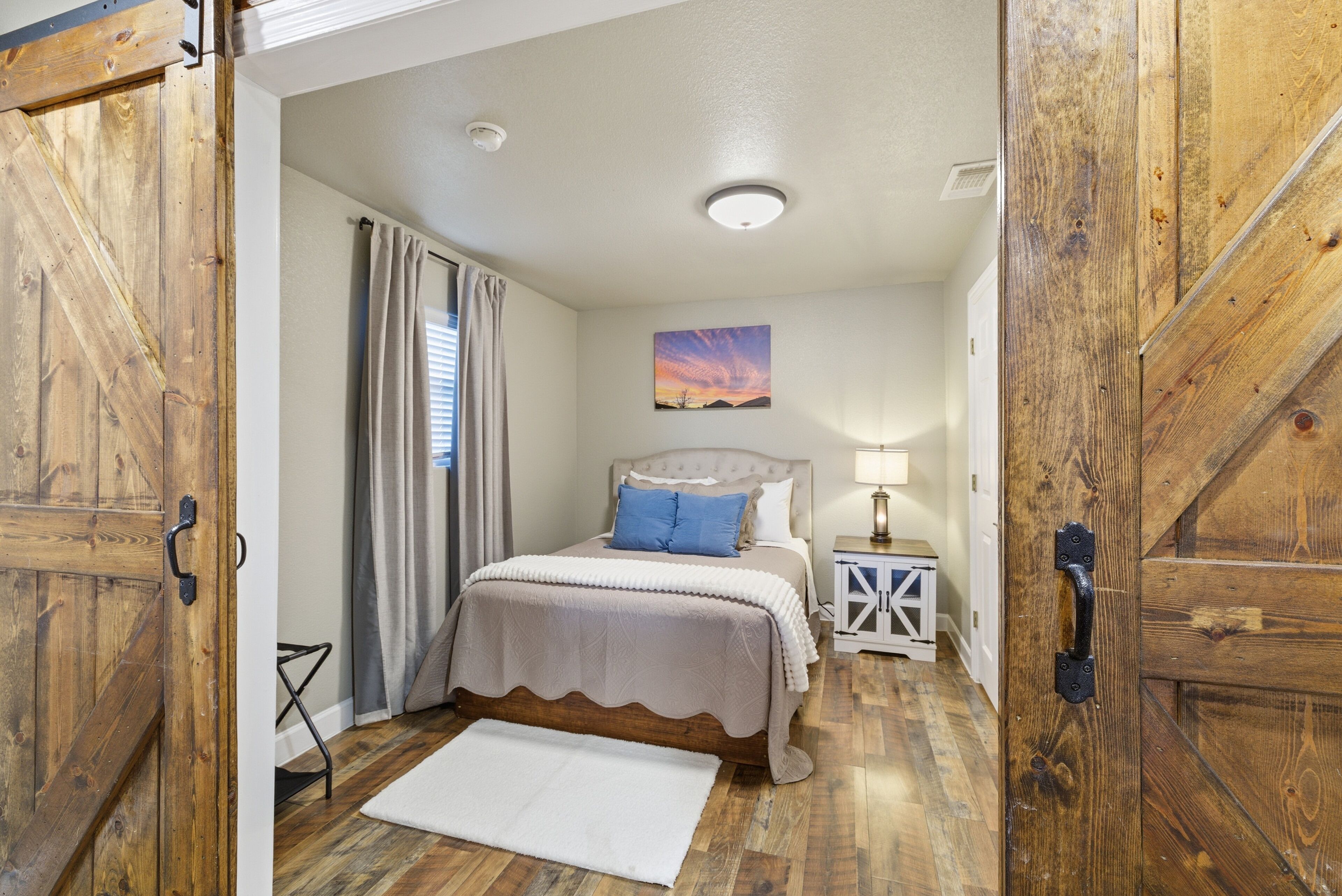 Un dormitorio con puerta de madera, una cama con colcha color crema y almohada azul, una mesita de noche con lámpara, y un cuadro en la pared.