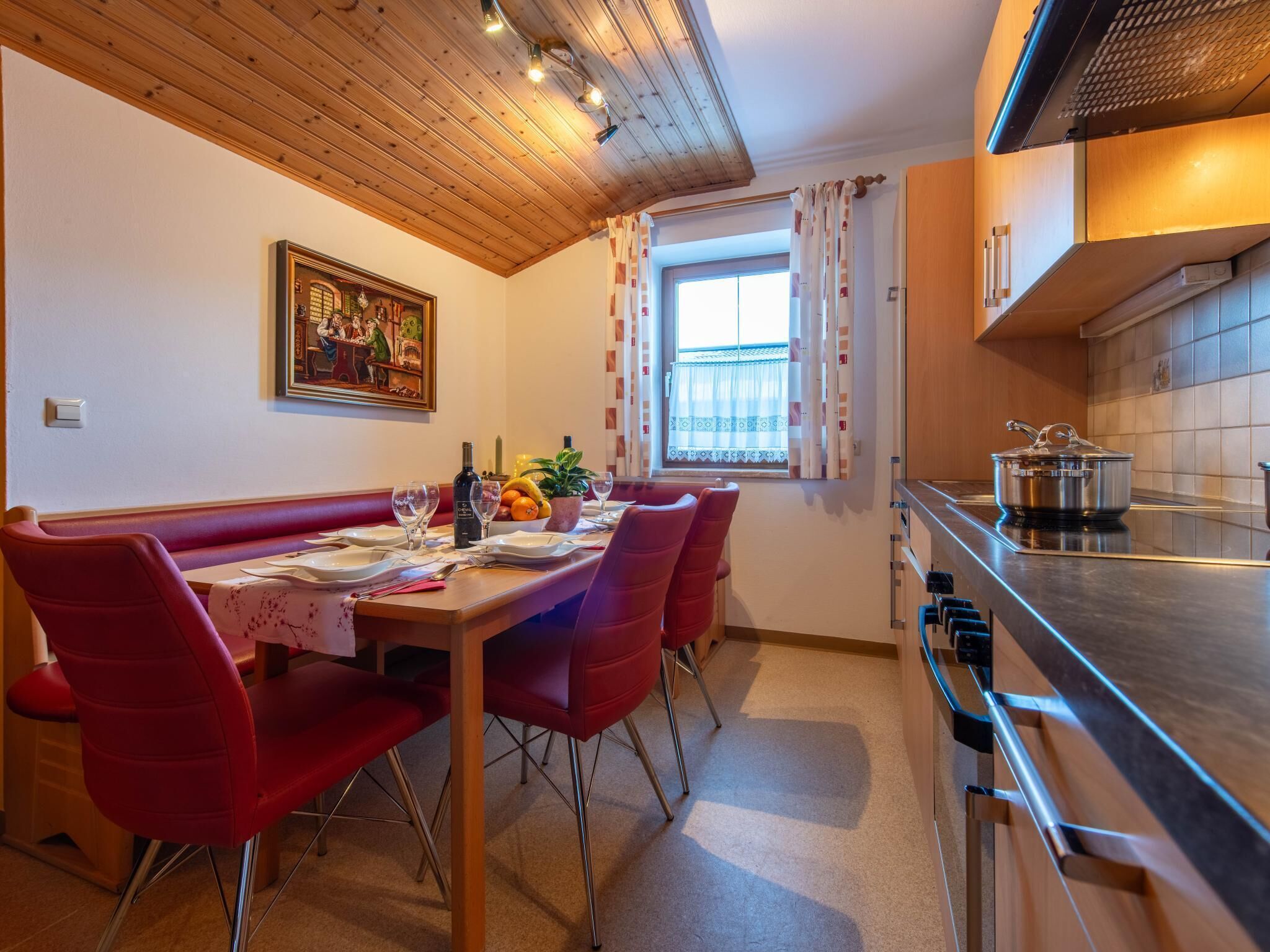A dining area with a wooden ceiling, a painting on the wall, a window with curtains, and a kitchen with a stove and pots.
