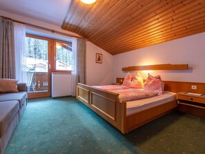 Ein gemütliches Schlafzimmer mit Holzbalkendecke, einem Bett mit blumigem Bettbezug, einem Nachttisch und Blick auf Bäume durch ein Fenster.