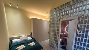 A bedroom with a bed, a wardrobe, a bathroom with a glass partition, and a small seating area with a cushion. - Atelier 12 - by Be Your Home (Milano)