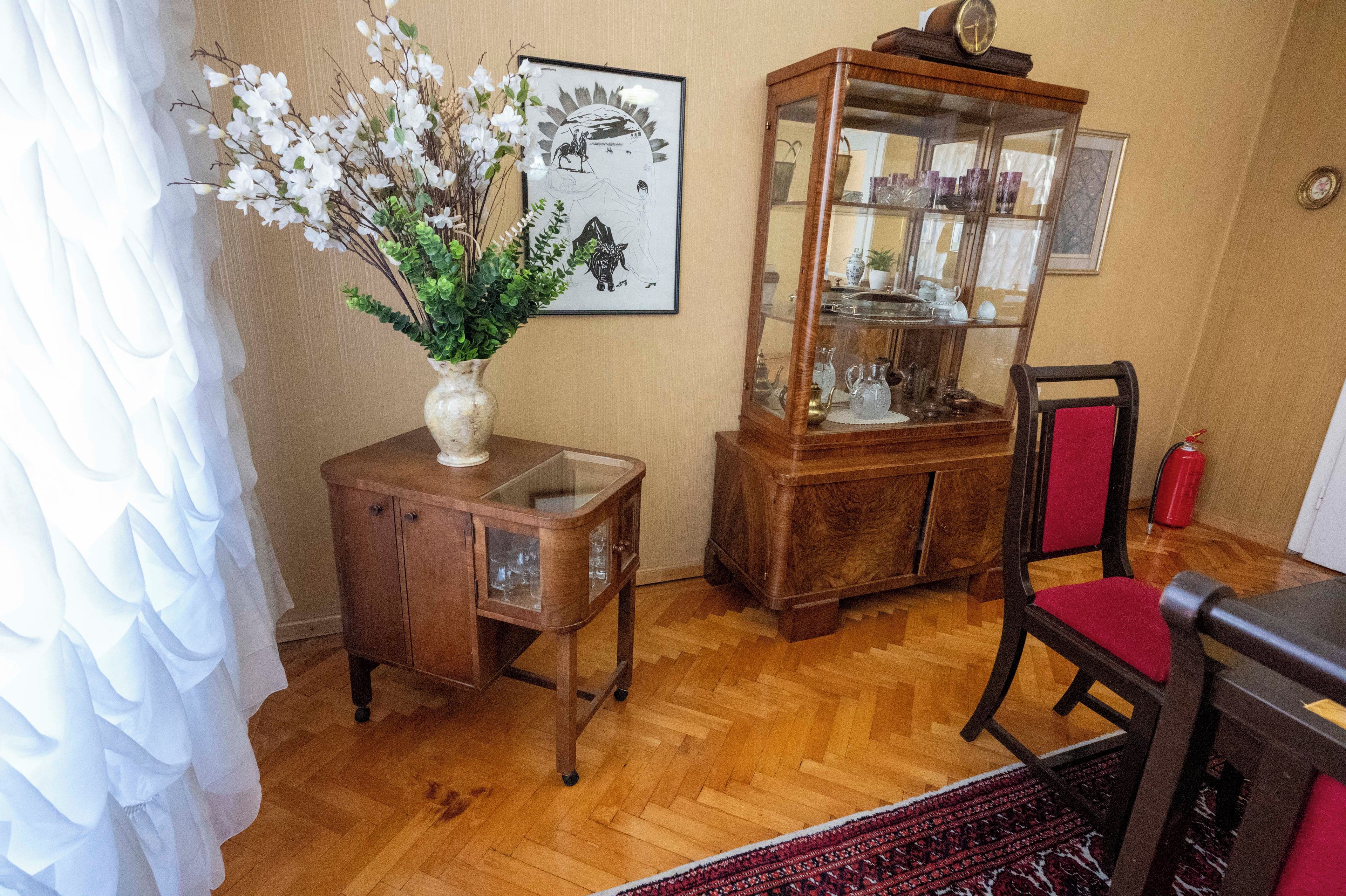 Ein Zimmer mit einem Holzkabinett, einer Glasvitrine, einer Vase mit Blumen und einem Stuhl.