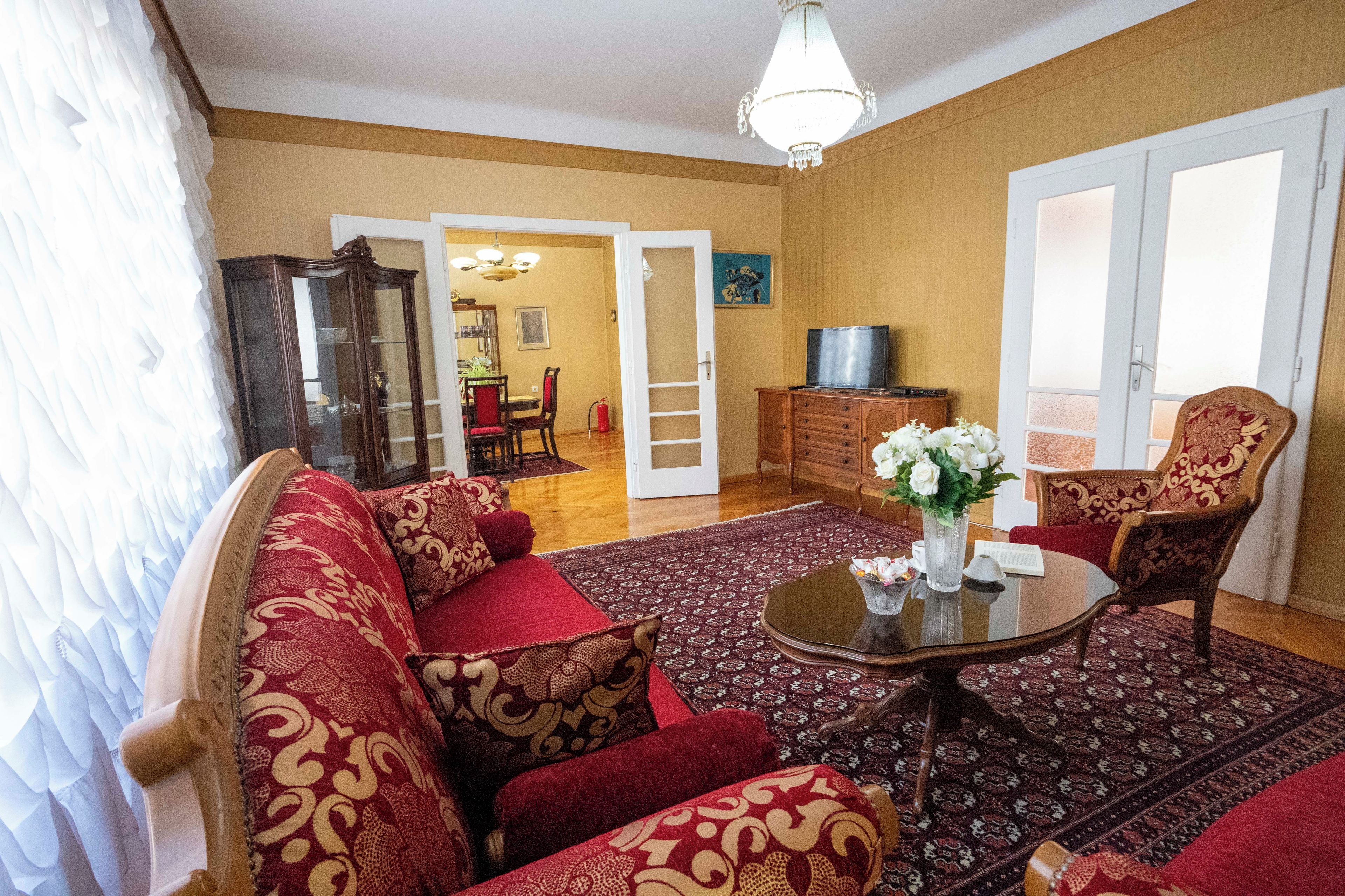 Ein Wohnzimmer mit einer Sofagarnitur, einem Couchtisch und einem gemusterten Teppich. Ein Fernseher steht auf einer Holzkommode und eine Tür führt in einen anderen Raum.