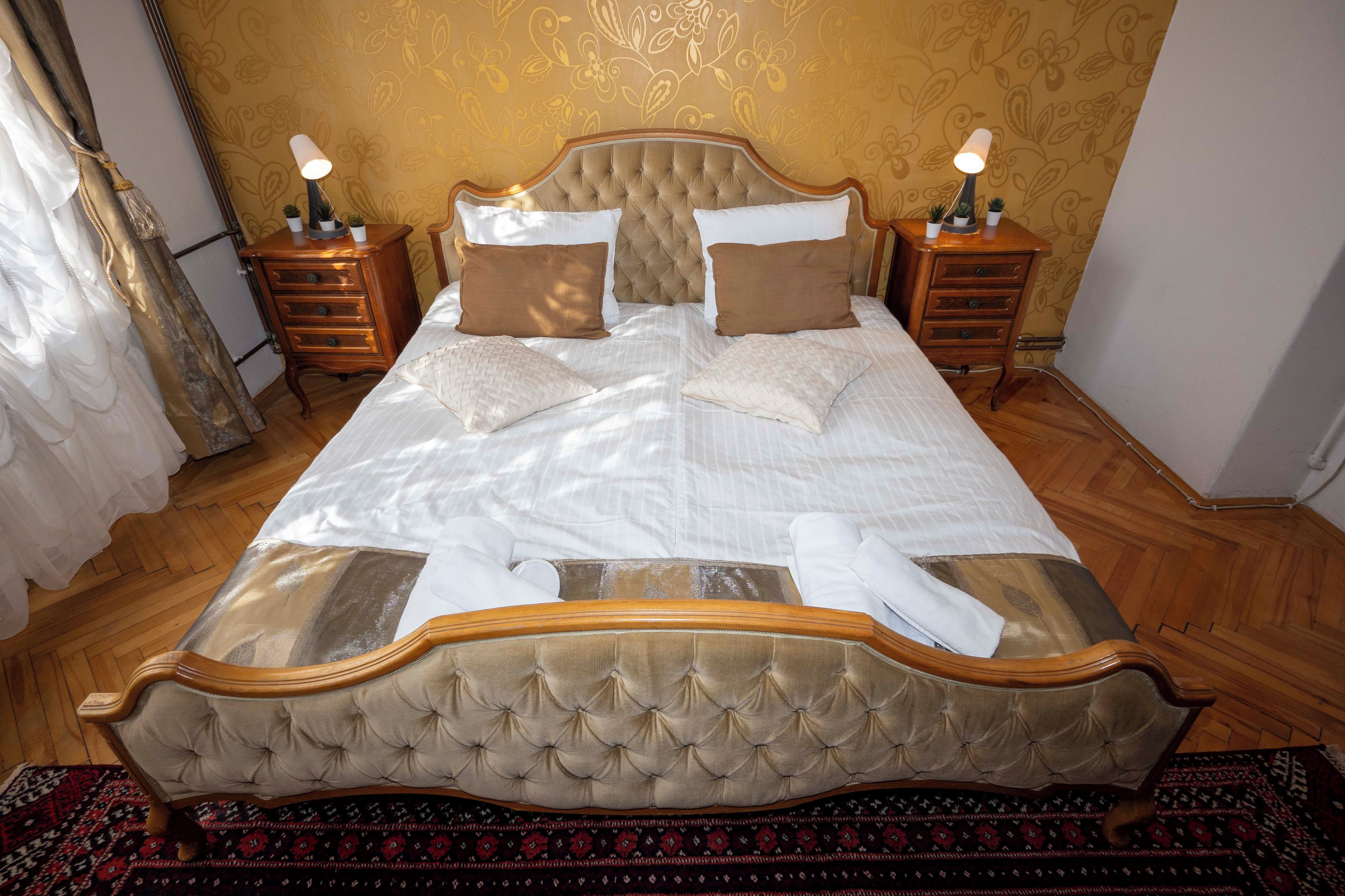 A large bed with a tufted headboard, two bedside tables with lamps, and a patterned carpet.