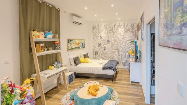 Un dormitorio con una cama, un escritorio, una estantería llena de juguetes y una pared decorada con motivos de hojas.