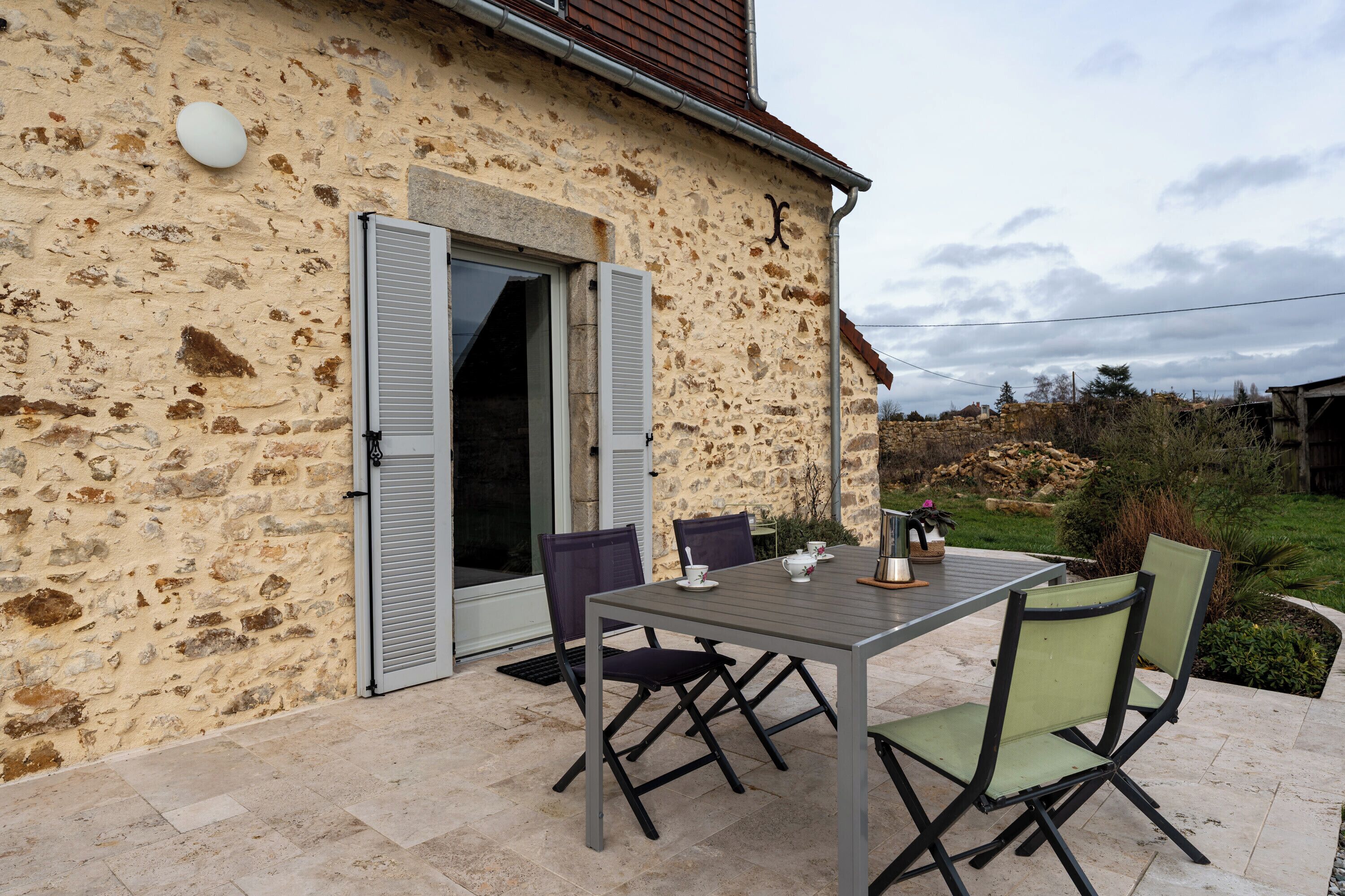 A stone building with a patio table and chairs, a door with shutters, and a small garden area.