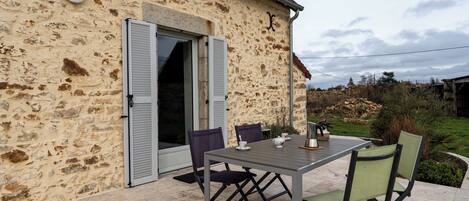 A stone building with a patio table and chairs, a door with shutters, and a small garden area.