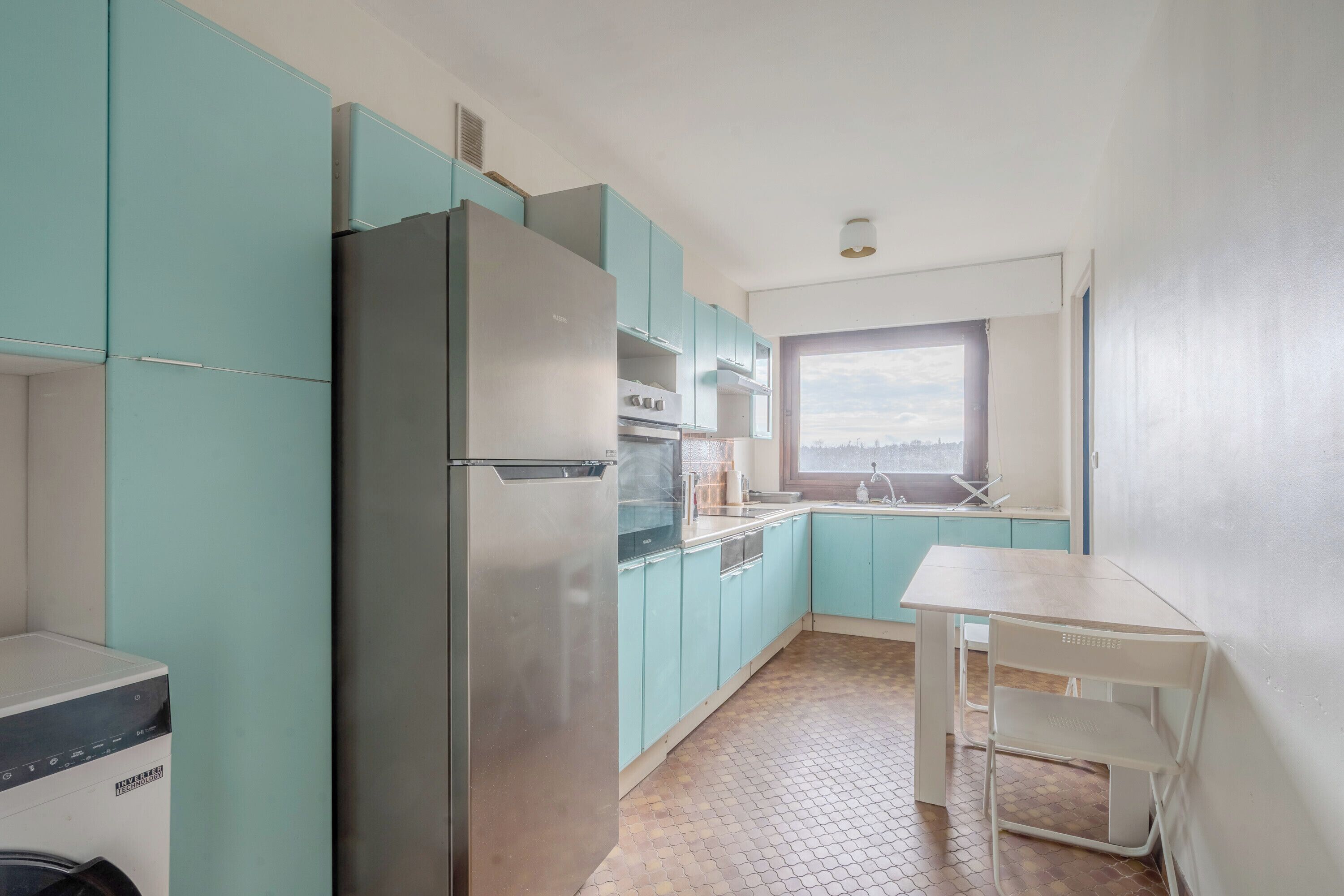 Een kleine keuken met blauwgroene kastjes, een roestvrijstalen koelkast, een magnetron en een spoelbak.