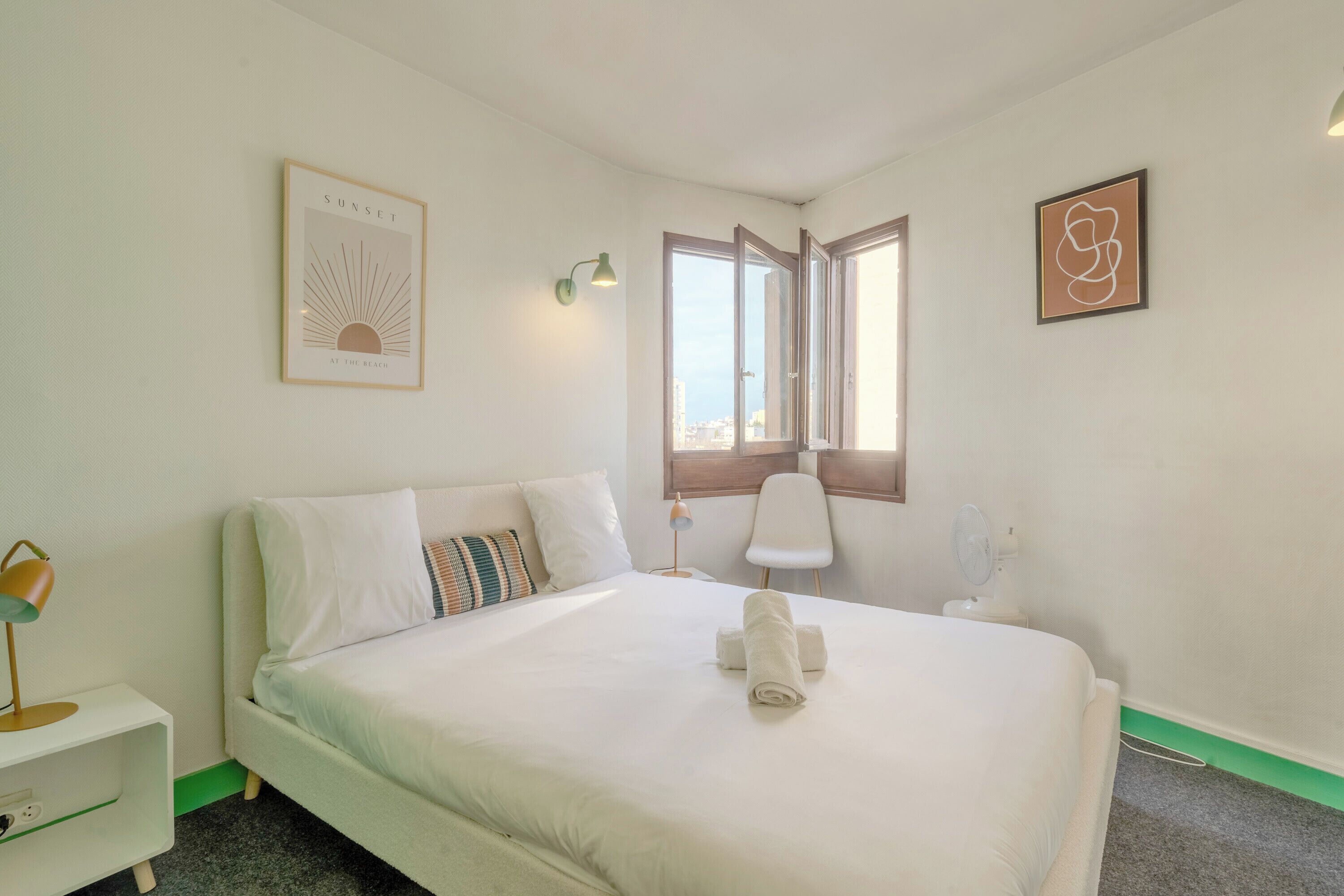 A neatly made bed with white bedding and a towel, a bedside table with a lamp, a chair, and a window with a view of buildings.