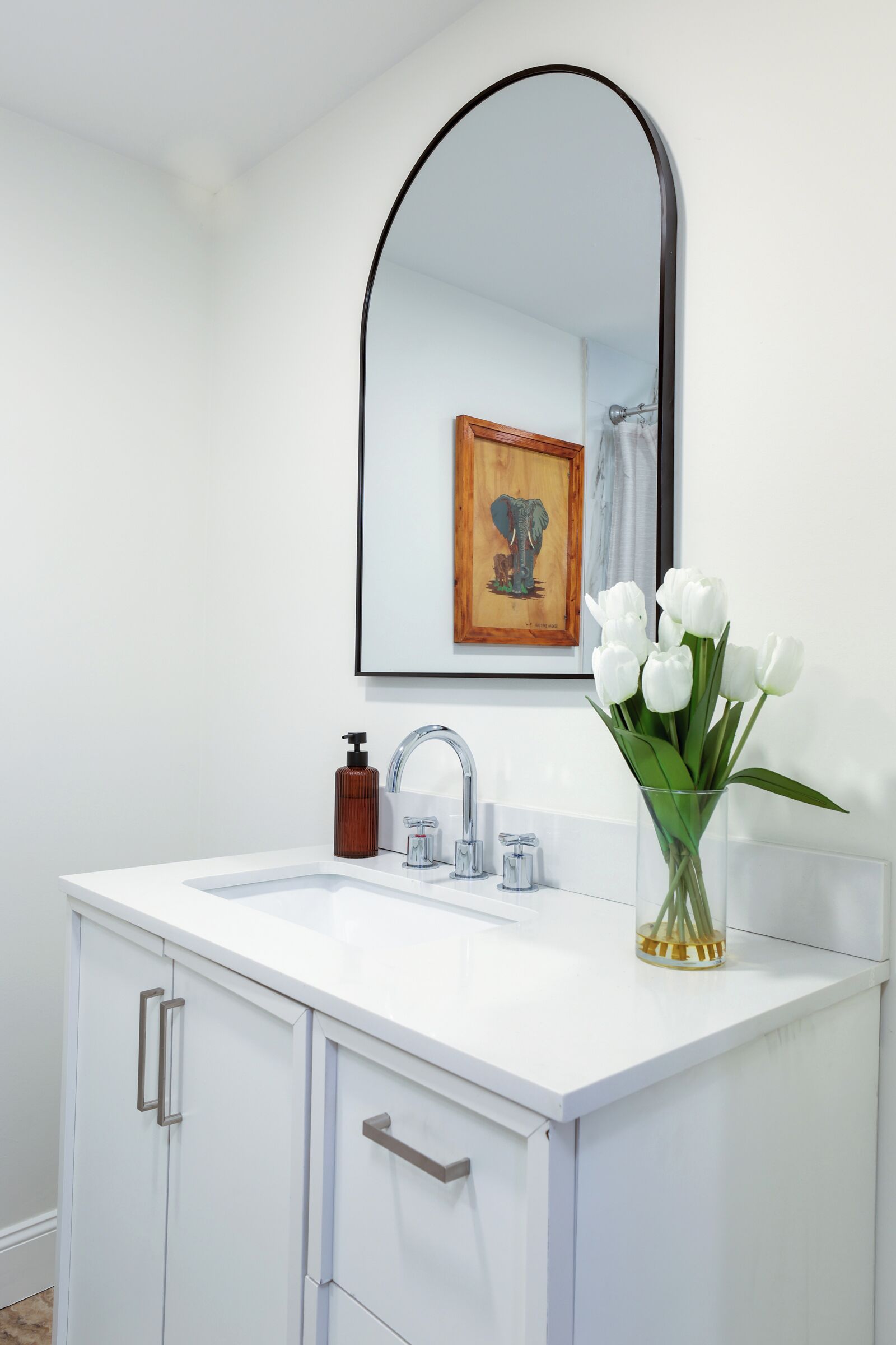 Une salle de bain avec un meuble-lavabo blanc, un miroir, un robinet, un distributeur de savon et un vase avec des fleurs blanches.