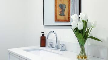 Une salle de bain avec un meuble-lavabo blanc, un miroir, un robinet, un distributeur de savon et un vase avec des fleurs blanches.