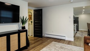 A modern living room with a flat-screen TV, a black entertainment unit, a potted plant, and a large door leading to another room. - Walkable 1BR in Village of Horseheads, Near I-86, Corning & Finger Lakes (Horseheads)