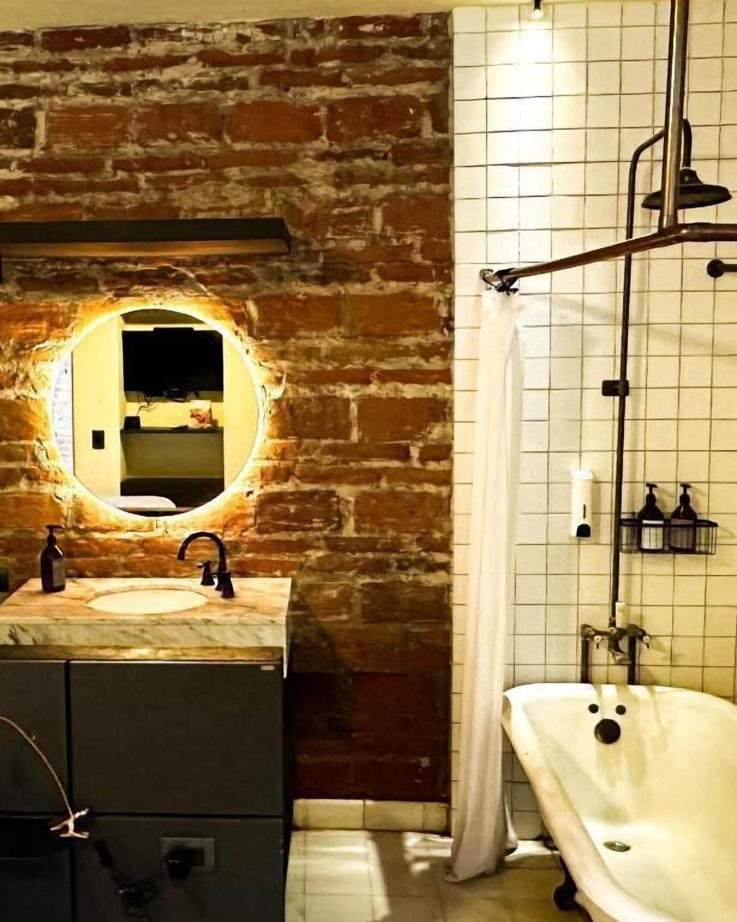 A bathroom with a stone wall, a round mirror, a sink, a bathtub, and a shower.