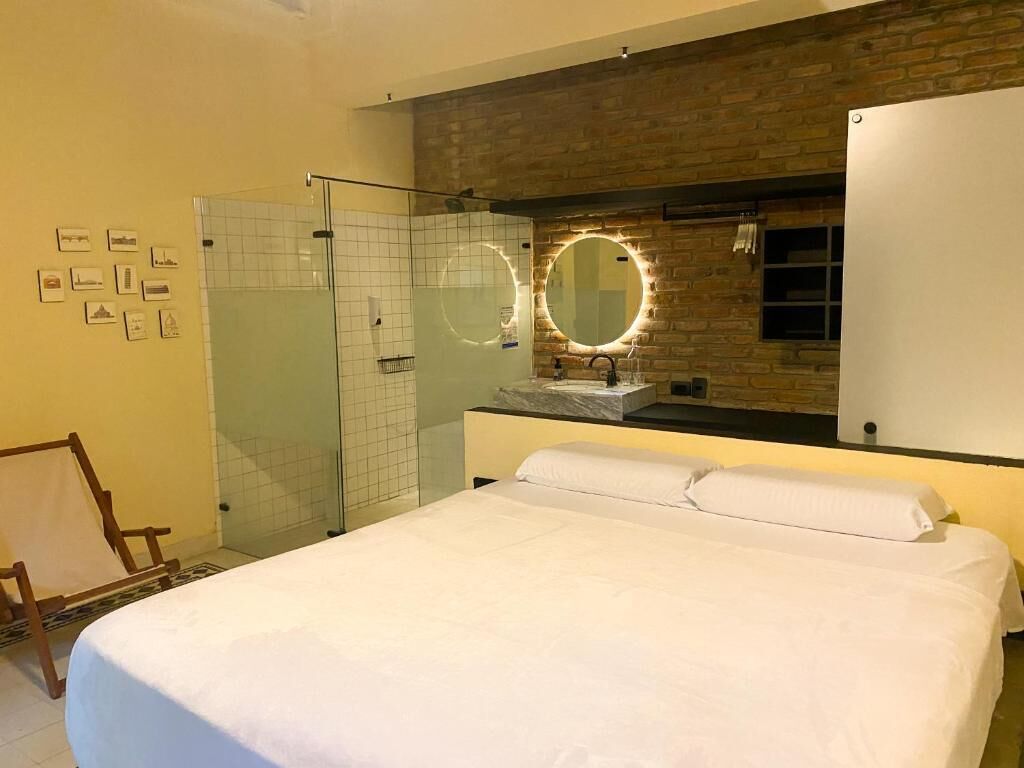 A hotel room with a large bed, a glass shower enclosure, a round mirror with lights, and a white wardrobe.