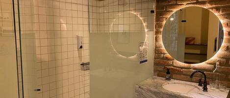 Un baño moderno con ducha de vidrio, lavamanos de mármol y espejo redondo con iluminación integrada.