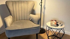 Un fauteuil beige à haut dossier, avec une lampe de sol noire placée à côté. Une table ronde, dotée d’un cadre métallique aux motifs géométriques, supporte un livre et une télécommande.