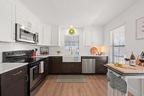 A modern kitchen with dark cabinets, a stainless steel microwave, a dishwasher, and a wooden countertop with a cutting board and wine glasses. - Blanco Mariposa - Hot Tub & Fire Pit (Fredericksburg)