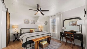 Ein Schlafzimmer mit einem Bett, Nachttischen, einem Schreibtisch, einem Stuhl, einem Deckenventilator und einem Fenster mit Vorhängen.