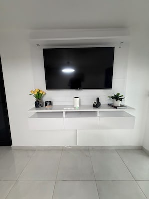 A modern living room with a large flat-screen TV mounted on a white wall, a white entertainment unit below, and a tiled floor.